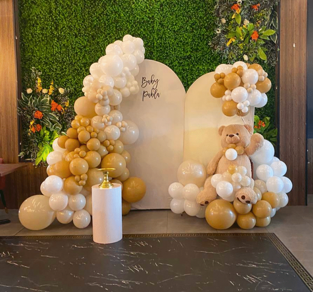 Balloon arch in beige and white tones with a large teddy bear and a baby shower backdrop decorated with greenery.